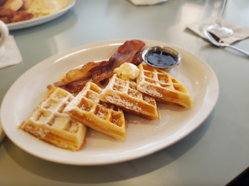 A plate of waffles topped with powdered sugar, butter, and syrup, accompanied by crispy bacon.