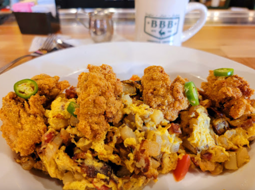 A plate of scrambled eggs with potatoes, peppers, and crispy fried chicken pieces, garnished with sliced jalapeños.
