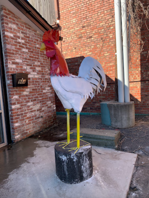 A large metal rooster sculpture stands on a concrete base outside a brick building.