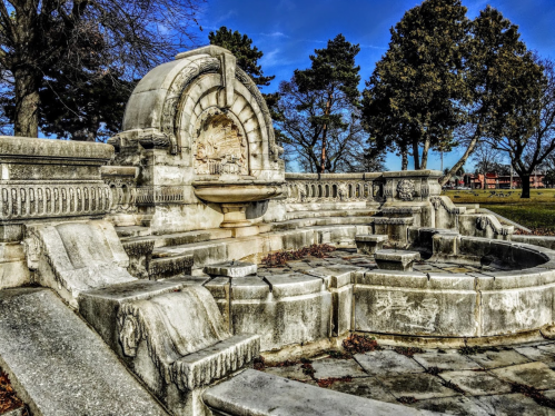 A historic stone fountain surrounded by trees, featuring intricate carvings and a clear blue sky.
