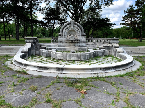 A weathered stone fountain surrounded by overgrown grass and trees in a park setting.