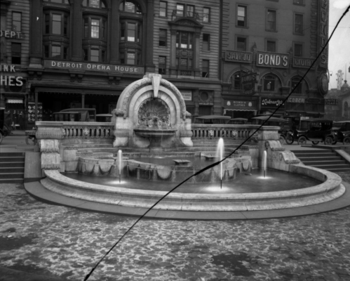 Historic fountain in a city square, surrounded by early 20th-century buildings and vintage cars.