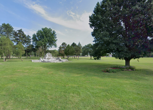 A spacious green park featuring a fountain, trees, and a clear blue sky.