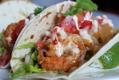 Two shrimp tacos filled with lettuce, diced tomatoes, and drizzled with sauce, garnished with cilantro and lime.