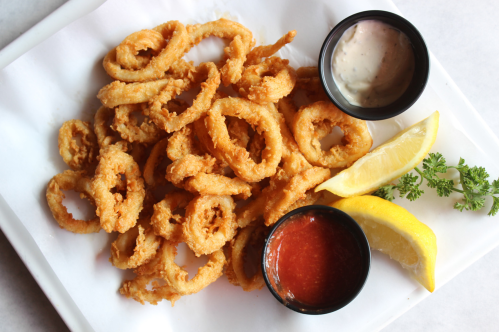 A plate of crispy fried calamari rings served with two dipping sauces and lemon wedges.