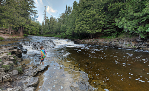 A child stands by a rocky riverbank, gazing at a waterfall surrounded by lush green trees.