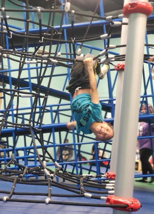 A child hangs upside down from a climbing structure, smiling while gripping the ropes in a play area.