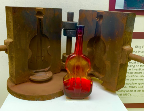 A glass violin bottle displayed between two metal molds in a museum setting.