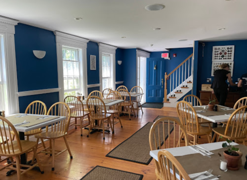 Cozy restaurant interior with blue walls, wooden tables, and chairs, featuring a staircase and large windows.