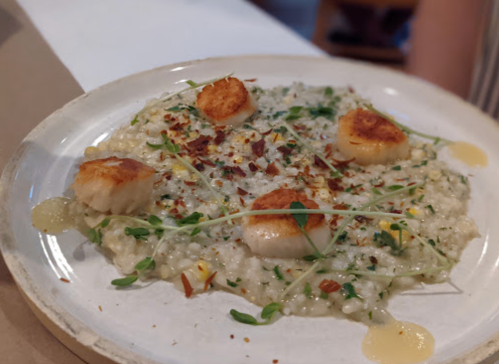 A plate of risotto topped with seared scallops, garnished with microgreens and a drizzle of sauce.