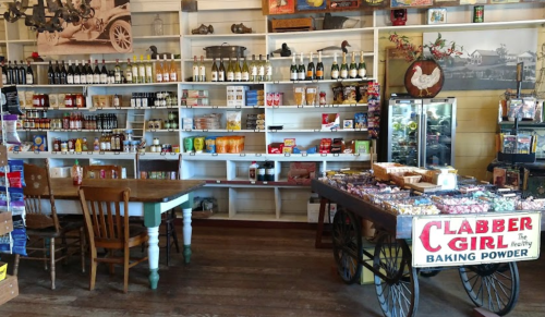 A rustic store interior featuring shelves of various products, a table, and a cart displaying Clabber Girl baking powder.