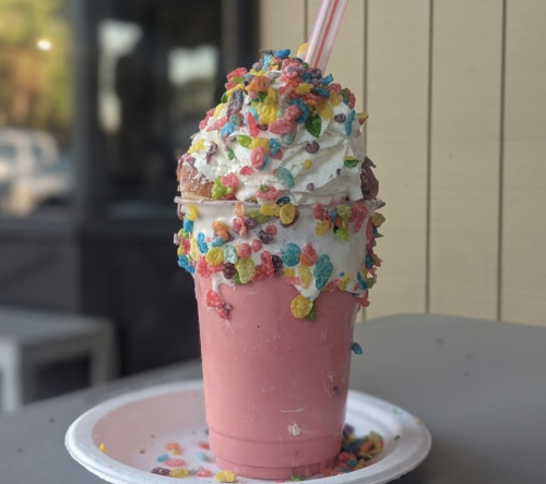 A colorful milkshake topped with whipped cream and sprinkled with fruity cereal, served in a clear cup with two straws.