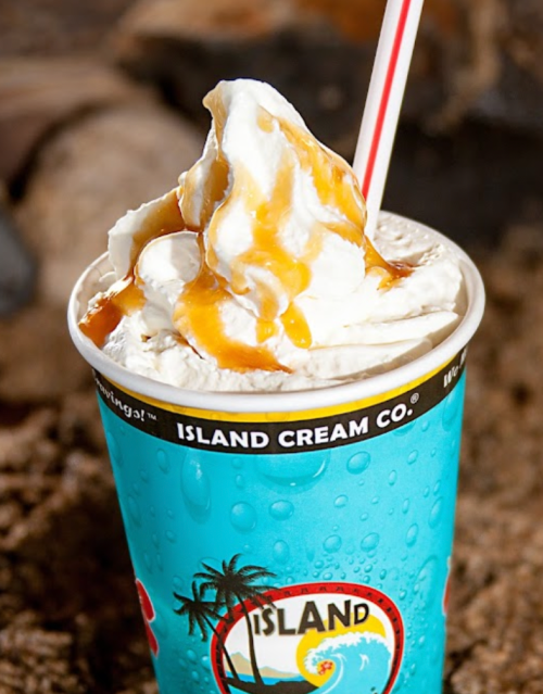 A blue cup filled with whipped cream and caramel sauce, set against a sandy background.