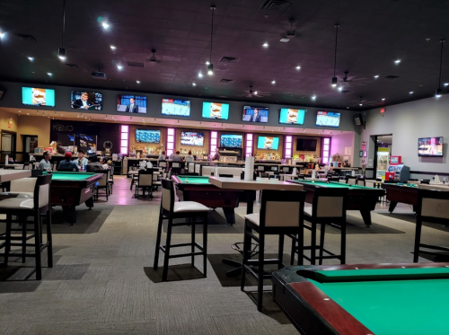 A spacious billiards hall with green pool tables, bar seating, and multiple screens displaying sports events.