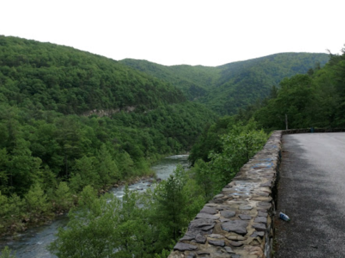 A scenic view of a river winding through lush green mountains, with a stone wall along the roadside.