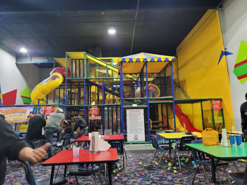 A colorful indoor play structure with slides and climbing areas, surrounded by tables and chairs in a lively setting.