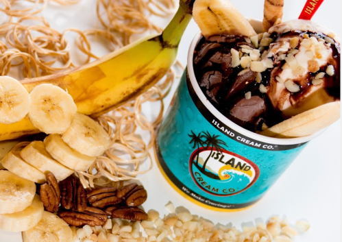 A cup of ice cream topped with chocolate sauce, bananas, and nuts, surrounded by banana slices and a whole banana.