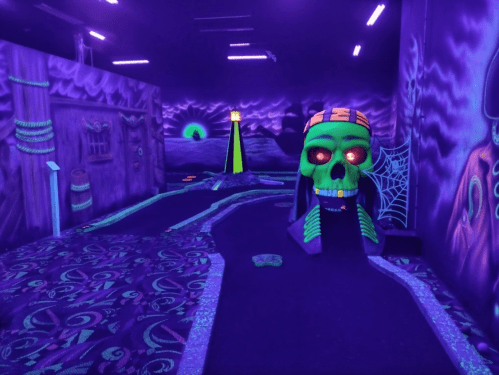 A glow-in-the-dark mini-golf course featuring a skull, spider webs, and eerie decorations in purple lighting.