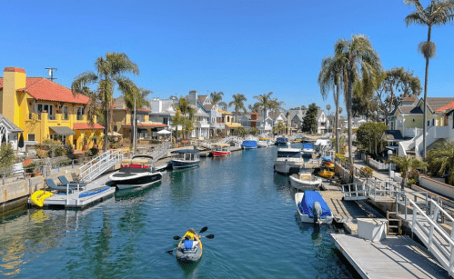 A sunny canal lined with colorful houses and boats, surrounded by palm trees and clear blue skies.