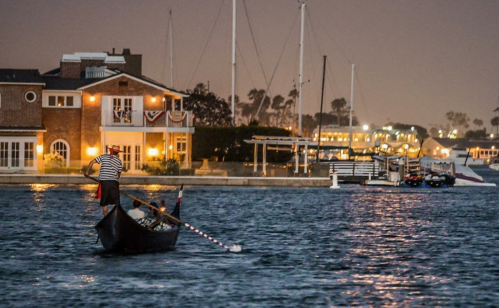 A gondolier rows a boat in a calm harbor at night, with illuminated buildings and sailboats in the background.