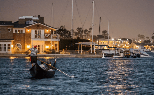 A gondolier rows a boat in a calm harbor at night, with illuminated buildings and sailboats in the background.