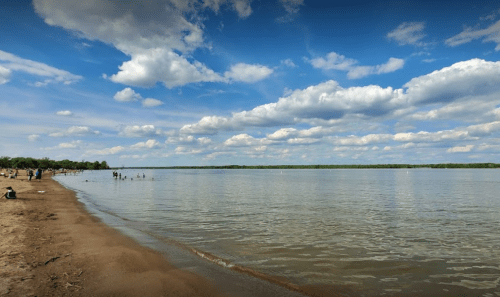 A serene beach scene with a sandy shore, calm water, and fluffy clouds in a blue sky. People enjoy the water in the distance.