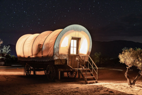 A lit-up covered wagon at night, surrounded by stars and desert landscape.