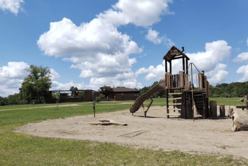 A wooden playground structure with a slide, set in a grassy area under a blue sky with fluffy clouds.