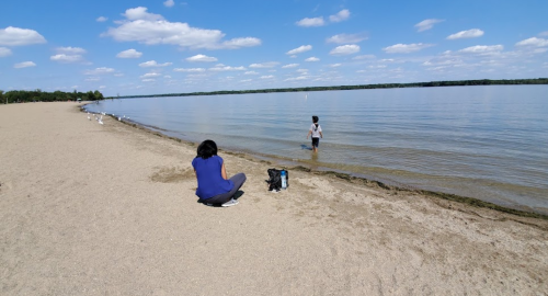 A person sits on the sandy beach while another stands in the water, with a calm lake and blue sky in the background.