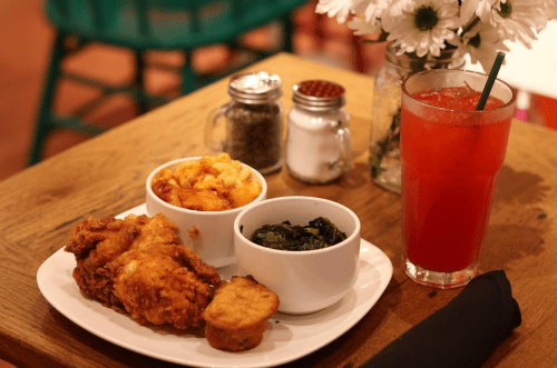 A plate of fried chicken, mac and cheese, collard greens, and a drink on a wooden table with flowers in the background.