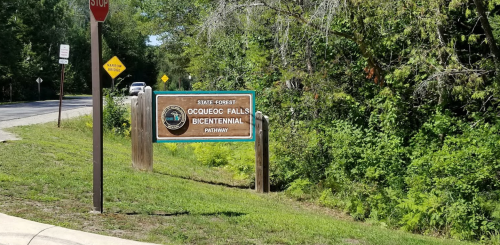 Sign for Ocuqueoc Falls Bicentennial Pathway in a wooded area, with a stop sign and road visible in the background.