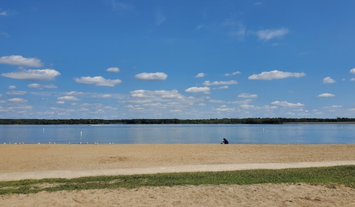 A serene lakeside view with a sandy beach, calm water, and fluffy clouds in a blue sky. A person sits by the shore.