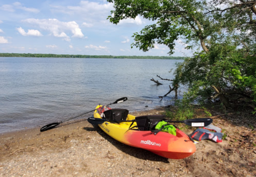 A colorful kayak rests on a sandy shore by a calm river, surrounded by trees and blue skies.