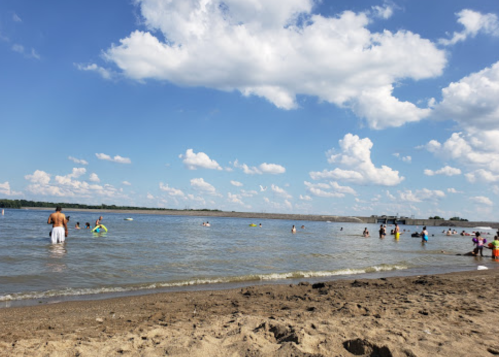 A sandy beach with people swimming and playing in the water under a blue sky with fluffy white clouds.