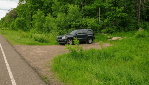 A black SUV parked on a gravel area beside a road, surrounded by lush green trees and grass.