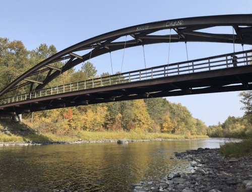 A curved metal bridge spans a calm river, surrounded by trees with autumn foliage.