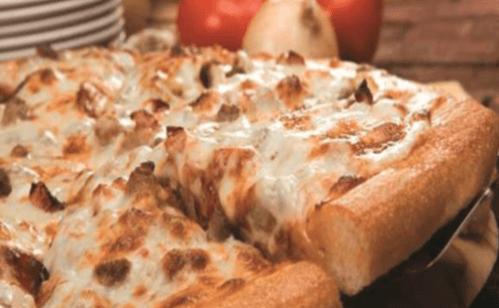 A close-up of a cheesy pizza with sausage, served on a wooden board, with tomatoes and onions in the background.