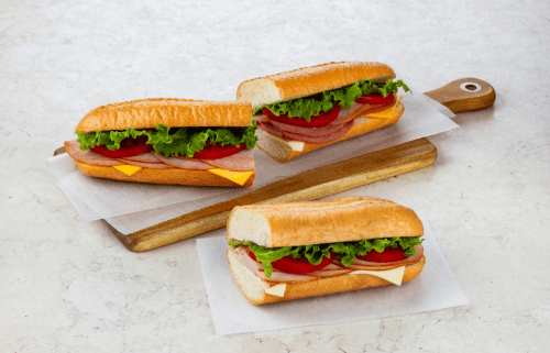 Three sandwiches on a wooden cutting board, filled with lettuce, tomato, cheese, and deli meats.