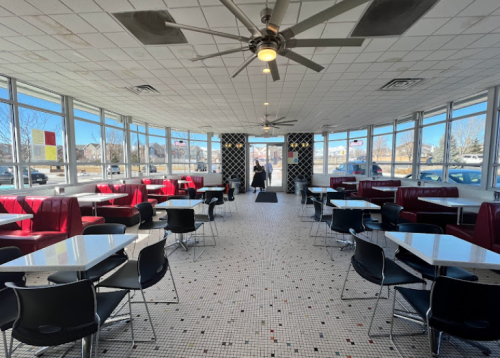 Bright diner interior with red booths, black chairs, and large windows, featuring a checkered floor and ceiling fans.