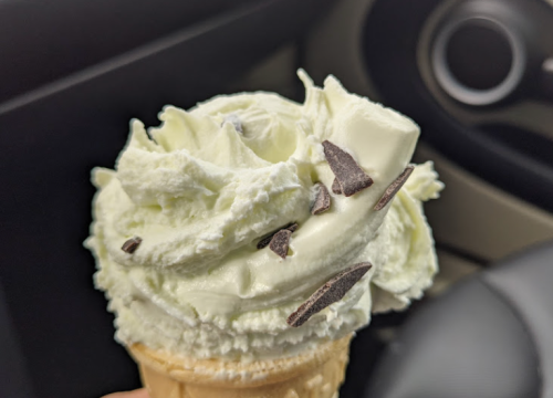 A close-up of a soft-serve ice cream cone topped with whipped cream and chocolate shavings, held inside a car.