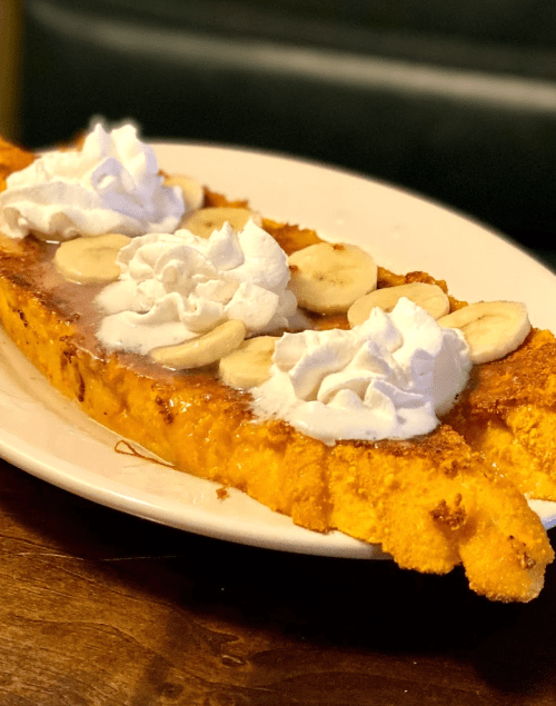 A large slice of French toast topped with banana slices and whipped cream, served on a white plate.