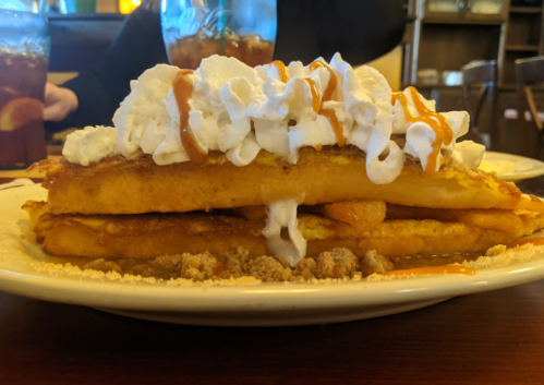 A stack of French toast topped with whipped cream and caramel sauce, served on a plate with crumbs underneath.