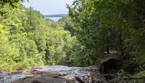 A scenic view from a rocky ledge overlooking a lush green forest and a distant lake.