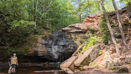 Two people stand near a rocky cliff with a small waterfall, surrounded by lush green trees and a calm pool of water.