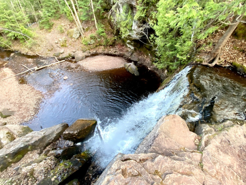 Aerial view of a waterfall cascading into a dark pool, surrounded by lush greenery and rocky terrain.