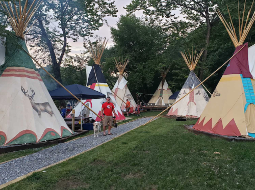 A man walks a dog among colorful tipis in a grassy area, surrounded by trees and a blue sky.