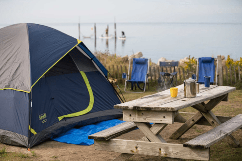 A camping scene with a tent, picnic table, and chairs by a calm lake, with paddleboarders in the background.