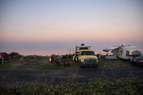 A campsite at dusk featuring a yellow truck, a camper, and a fire pit with a scenic view in the background.