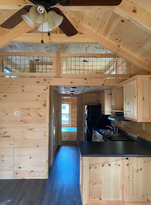 Cozy wooden interior of a tiny house featuring a kitchen, ceiling fan, and loft area with a railing.