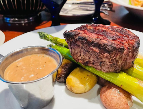 A perfectly cooked steak atop grilled asparagus and potatoes, served with a side of sauce in a small metal cup.